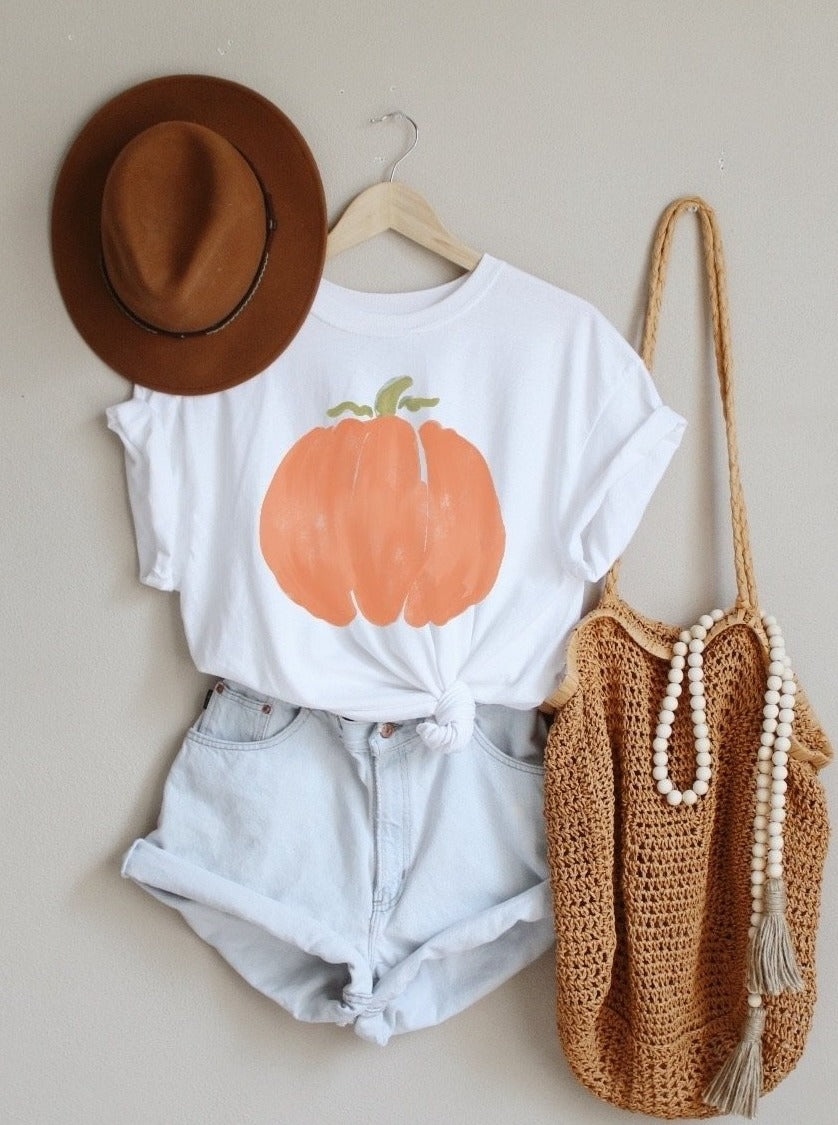 Watercolor Orange pumpkin tee hanging on the wall with boho straw purse, paired with cutoff jean shorts, and brown felt hat.